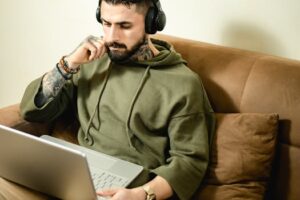 Tattooed man in hoodie with headphones working on laptop from home, embodying modern remote work lifestyle.