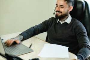 A young tattooed man in a headset works at a desk with a laptop and documents.