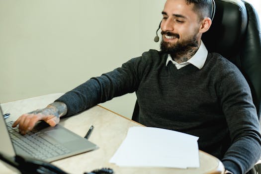 A young tattooed man in a headset works at a desk with a laptop and documents.
