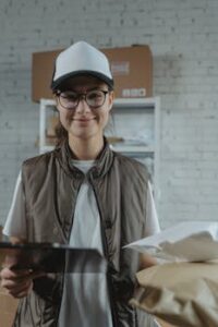 A young female courier smiling while managing packages with a tablet in a warehouse environment.