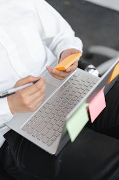 Adult wearing white long sleeves working on a laptop and holding sticky notes, showcasing multitasking.
