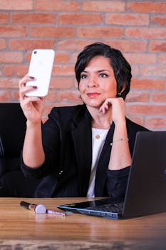 Confident woman taking a selfie with her smartphone in a stylish, brick-walled office.