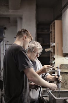 Side view of elderly foreman in eyeglasses and assisting trainee handling detail in workshop