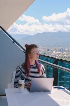 Young woman working on a laptop on a balcony with a scenic city view.