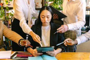 Young woman working on a tablet, surrounded by colleagues seeking attention in a busy office setting.